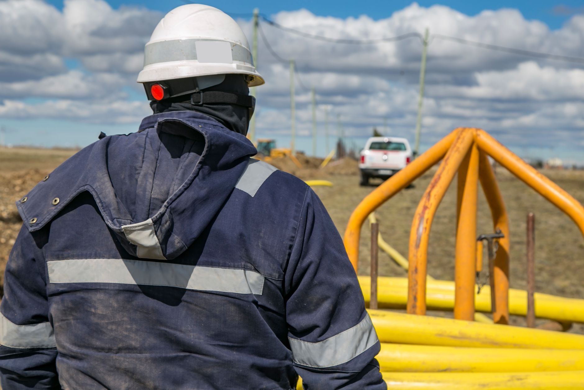 A pipeline technician in a hard hat and reflective jacket inspecting a yellow above-ground riser in an open field